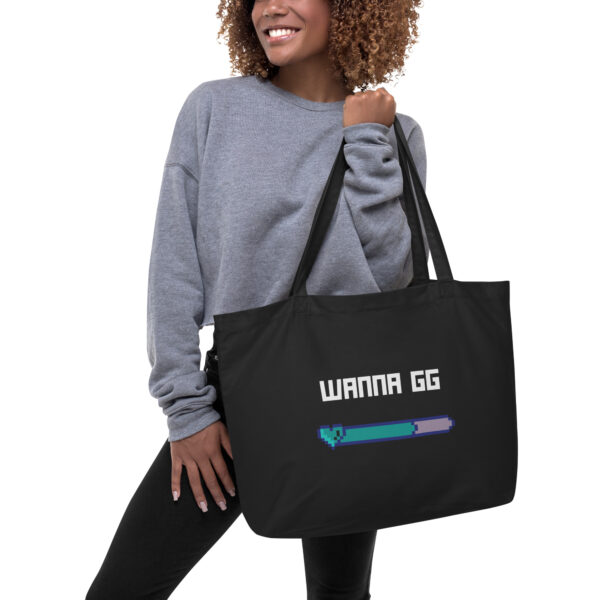 A woman holding a black tote bag on which is written "wanna gg" in white with a green and blue lifebar underneath