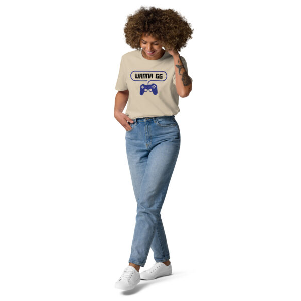 A woman posing head-on wearing a beige tshirt on which is writting "wanna gg" in black with a blue PlayStation controller