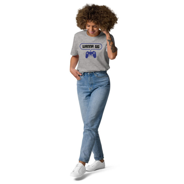 A woman posing head-on wearing a grey tshirt on which is writting "wanna gg" in black with a blue PlayStation controller